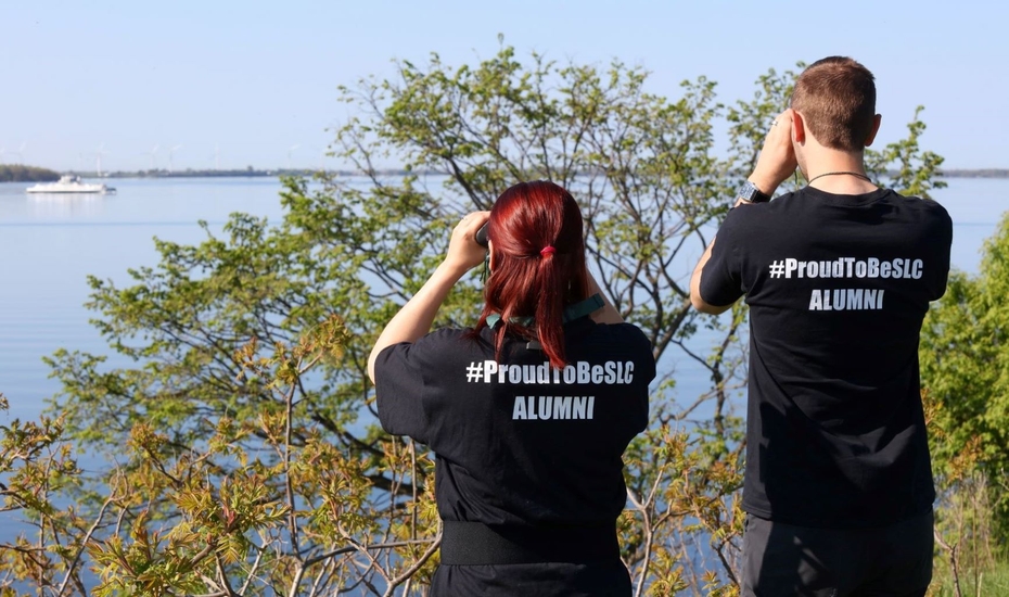 Two people hold binoculars up to their eyes and stare out at a boat on the water. They are both wearing t-shirts that say Proud to Be SLC alumni